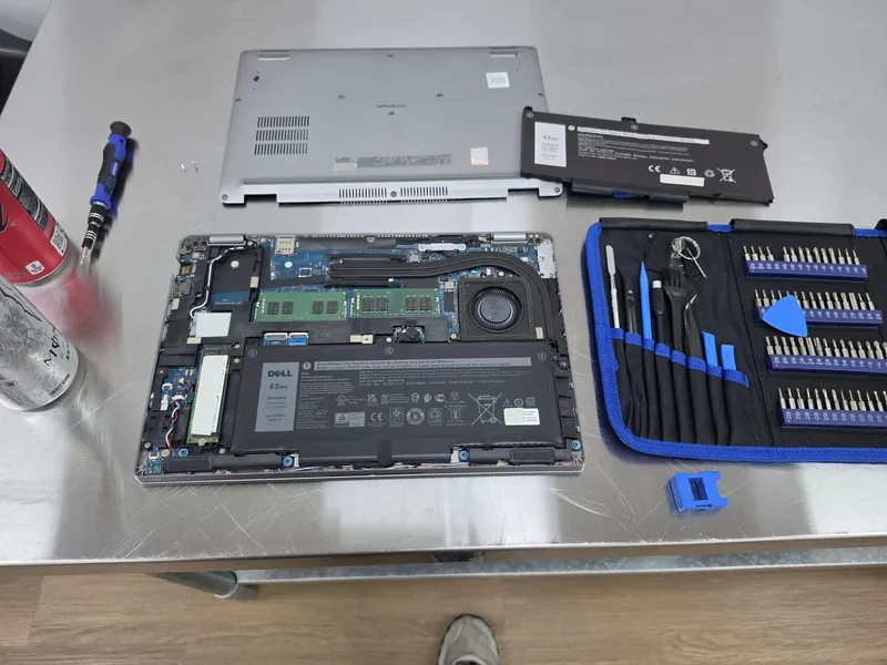 An opened Dell Latitude laptop on a workbench beside its removed battery and a tool kit.